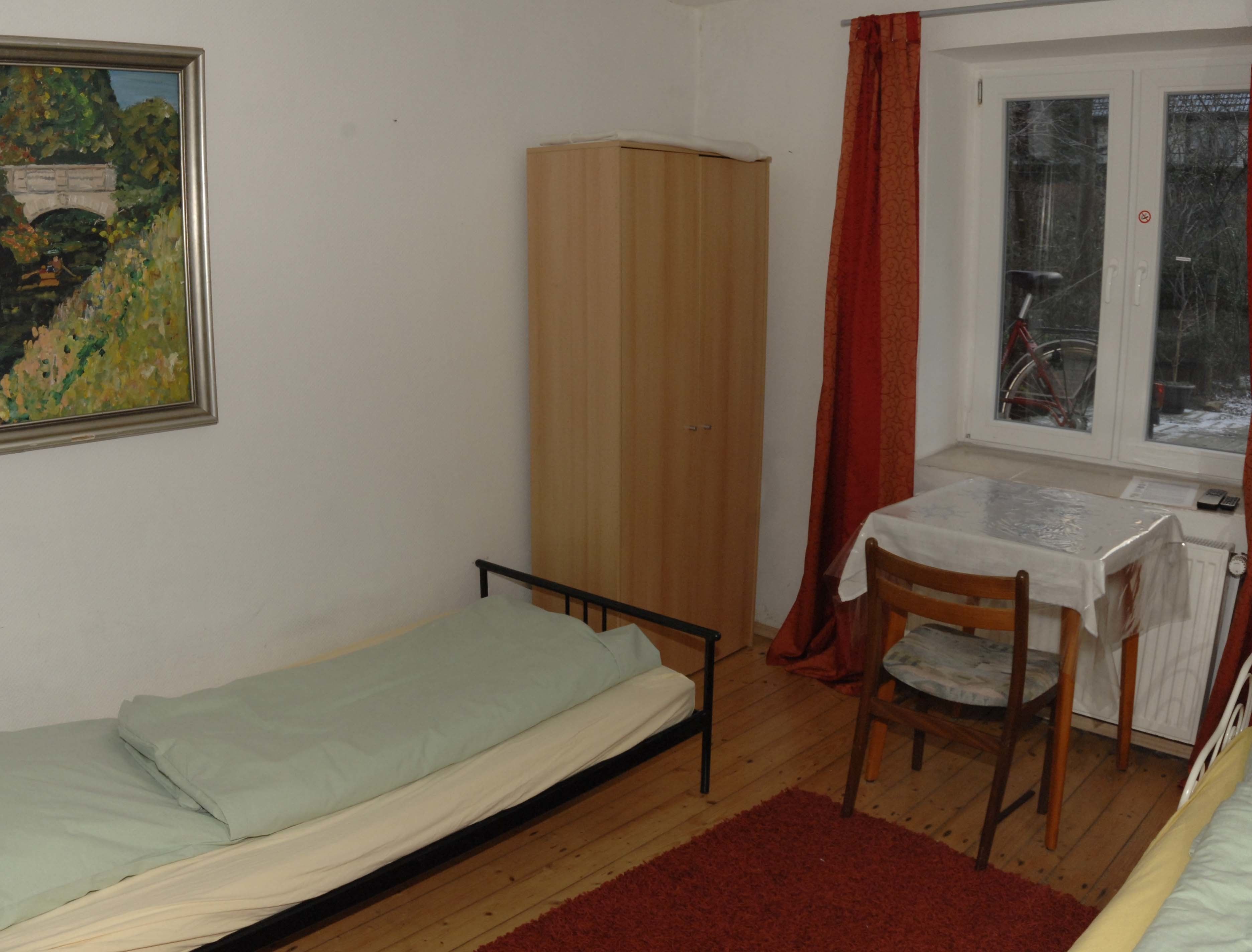 A small room with a single bed, a wooden wardrobe, a table with a chair, and a window with a view of a bicycle outside.