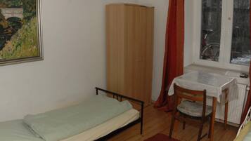 A small room with a single bed, a wooden wardrobe, a table with a chair, and a window with a view of a bicycle outside.