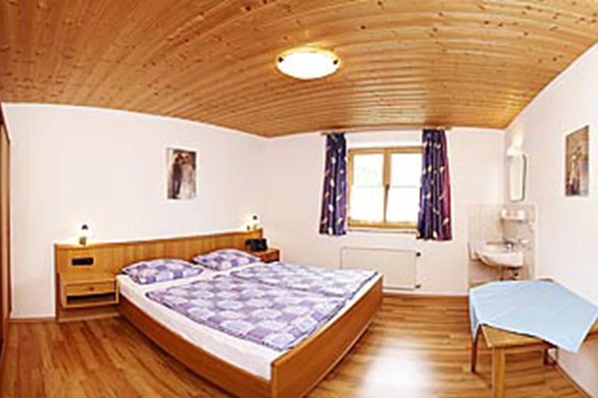 A bedroom with a wooden ceiling, a bed with a purple and white bedspread, a wooden nightstand, a window with curtains, a bedside table, and a small desk with a chair.