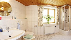 A bathroom with a wooden ceiling, a large sink, a toilet, a shower, and a window with curtains. - Beim Draxl (Reit im Winkl)
