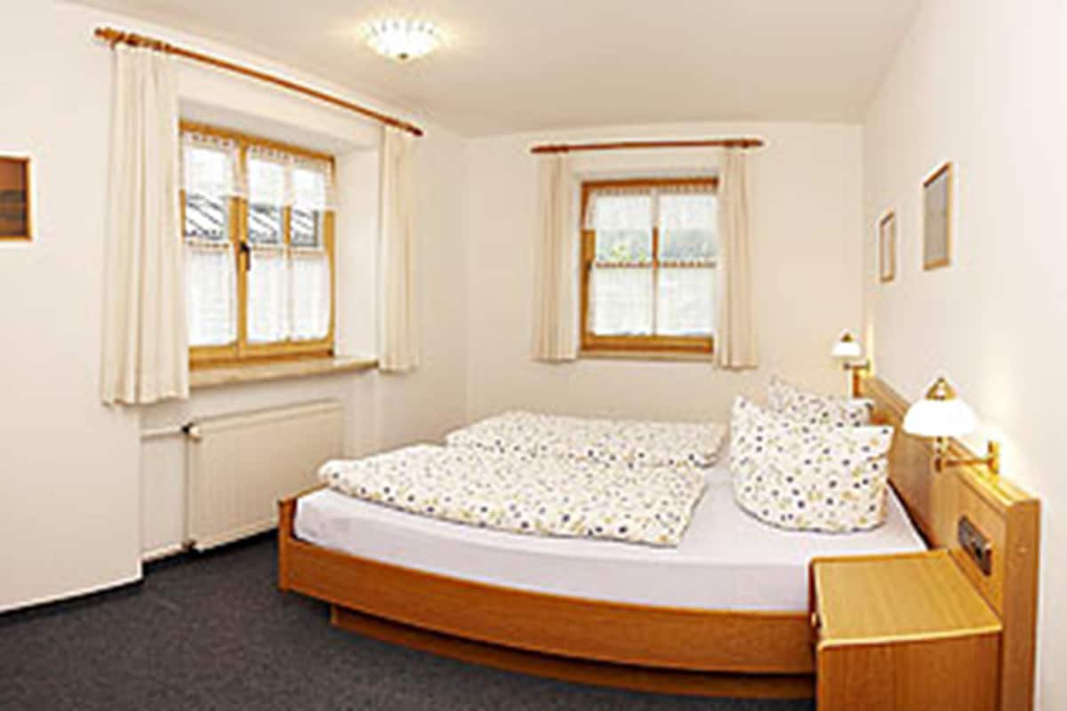 A bedroom with a single bed, two windows with curtains, a bedside table with a lamp, and a radiator.