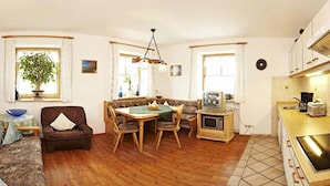 A cozy living room with a sofa, dining table, and chairs, a kitchen with a stove and microwave, and a large window with a plant. - Beim Draxl (Reit im Winkl)
