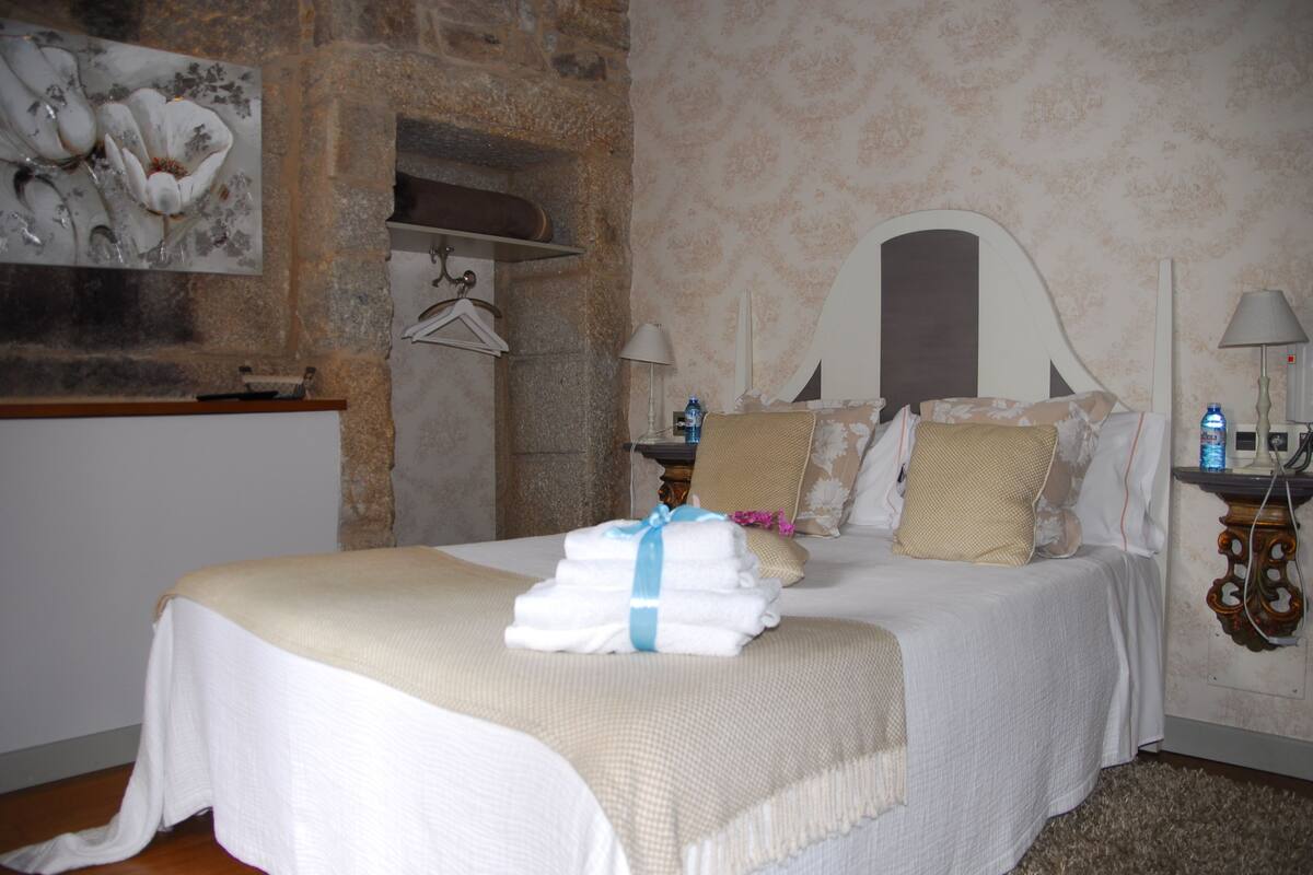 A neatly made bed with a folded towel and a blanket, a wooden nightstand with a lamp, a wall-mounted towel rack, and a painting of flowers.
