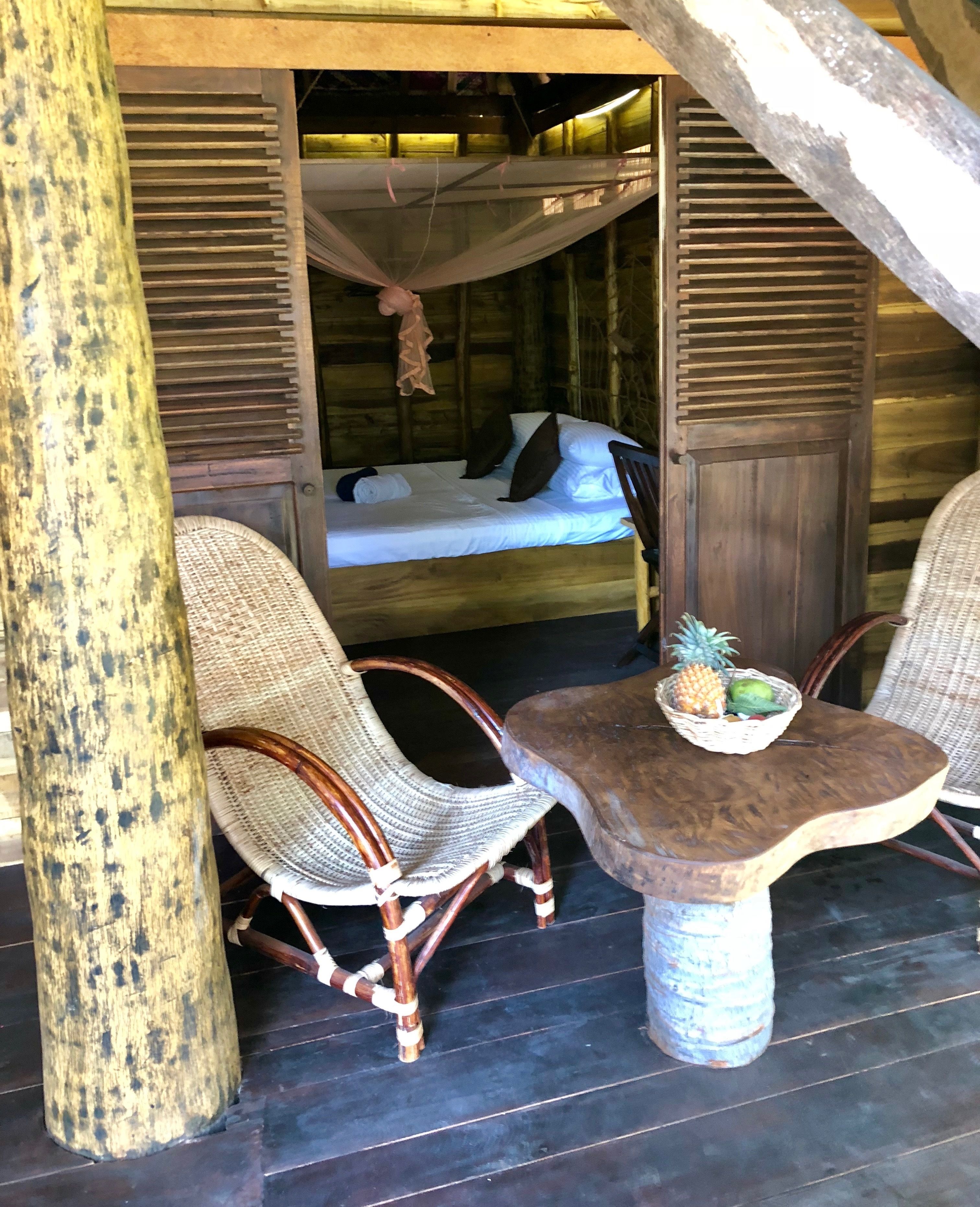 Una habitación rústica de madera con una cama, sillas de mimbre y una mesa con cesta de frutas.