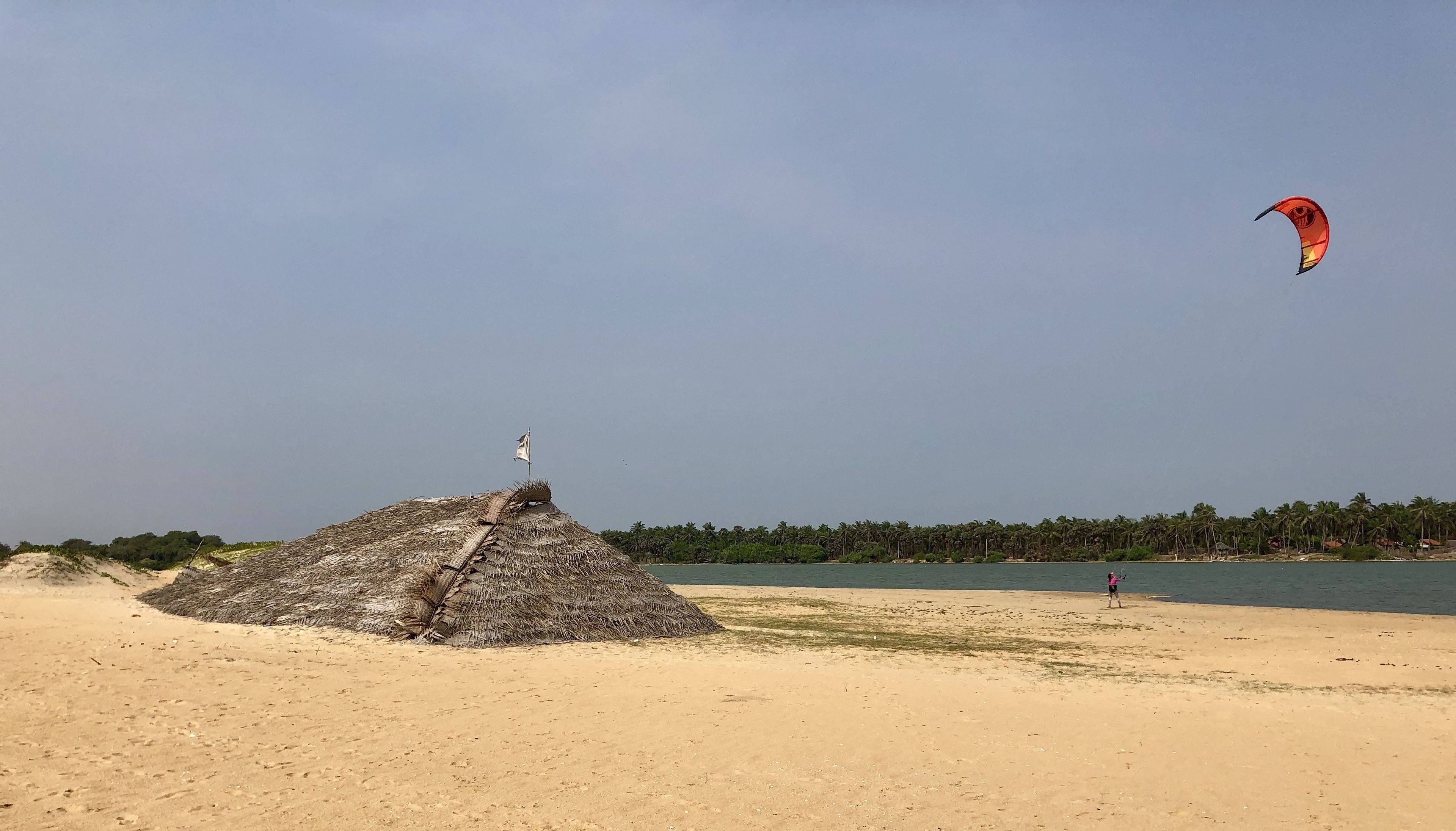 Una playa arenosa con una estructura de techo de paja y una bandera en la parte superior, una masa de agua y una persona volando una cometa.