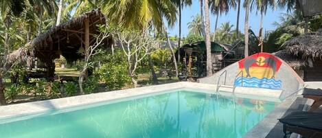 A swimming pool with a mural of dolphins, surrounded by palm trees and a thatched-roof structure.