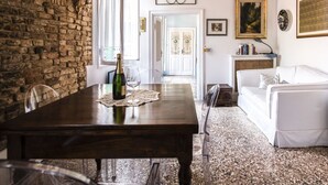 A dining area with a wooden table, chairs, and a bottle of wine. There is a white sofa and a small cabinet in the background. - Venice Giardini Biennale Apartment (Venice)