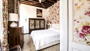 A bedroom with a wooden ceiling, a bed with a white bedspread, a wooden dresser, and floral wallpaper. - Venice Giardini Biennale Apartment (Venice)