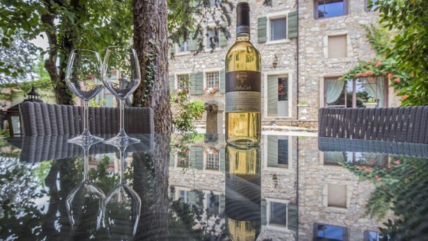 Una botella de vino y dos copas sobre una superficie reflectante, con un edificio de piedra al fondo.