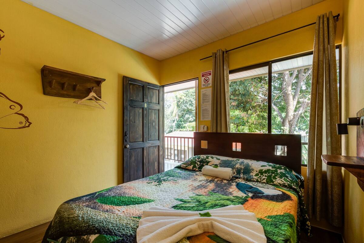 A room with a bed, a wooden headboard, a window with curtains, and a view of trees outside.