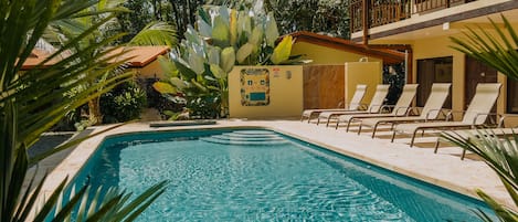 Ein Swimmingpool mit Liegestühlen, umgeben von tropischer Vegetation und einem Gebäude mit Balkon.
