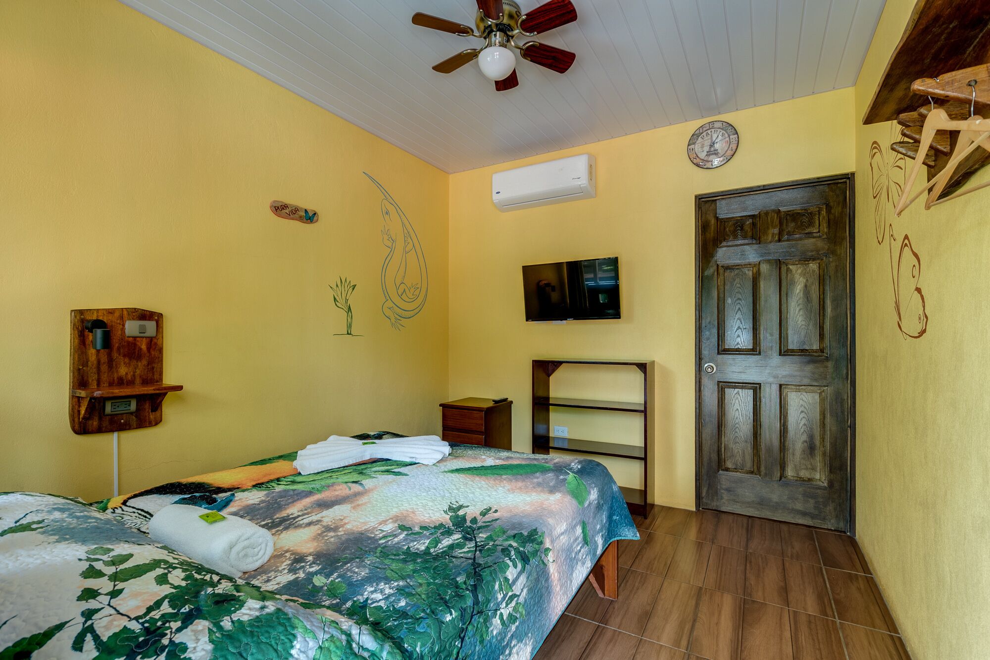 Une chambre à coucher avec un lit, un ventilateur de plafond, un téléviseur fixé au mur, une porte en bois et une petite étagère.
