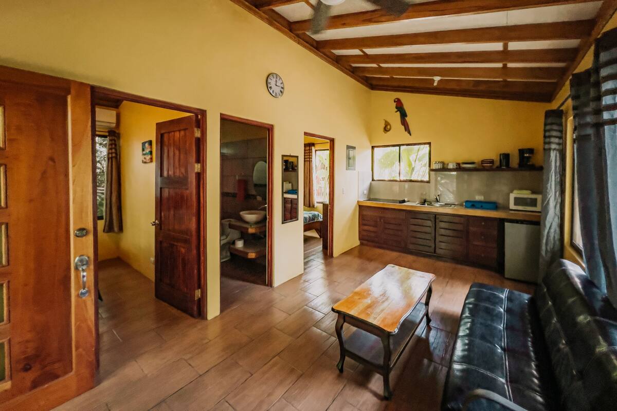 A spacious living area with a wooden floor, a dining table, a sofa, and a kitchen area with a microwave and cabinets.
