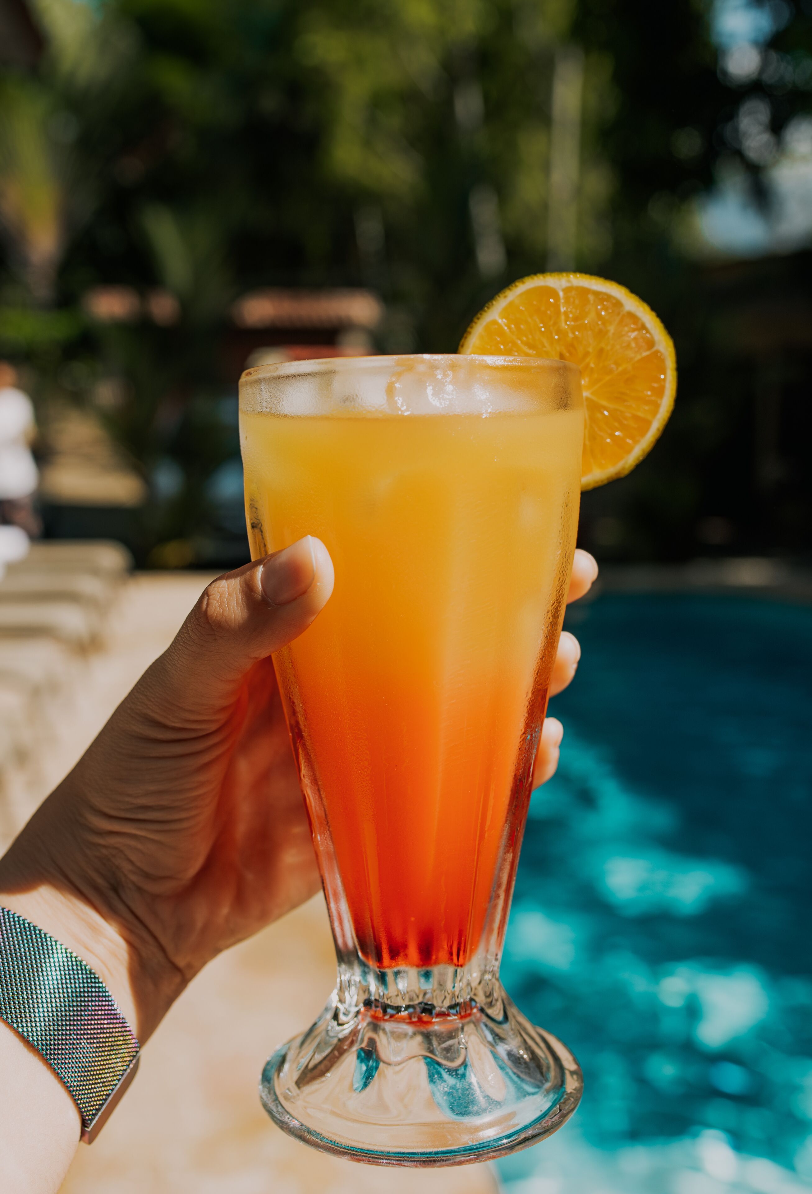 A hand holding a tall glass with a gradient orange drink, garnished with an orange slice, near a pool.