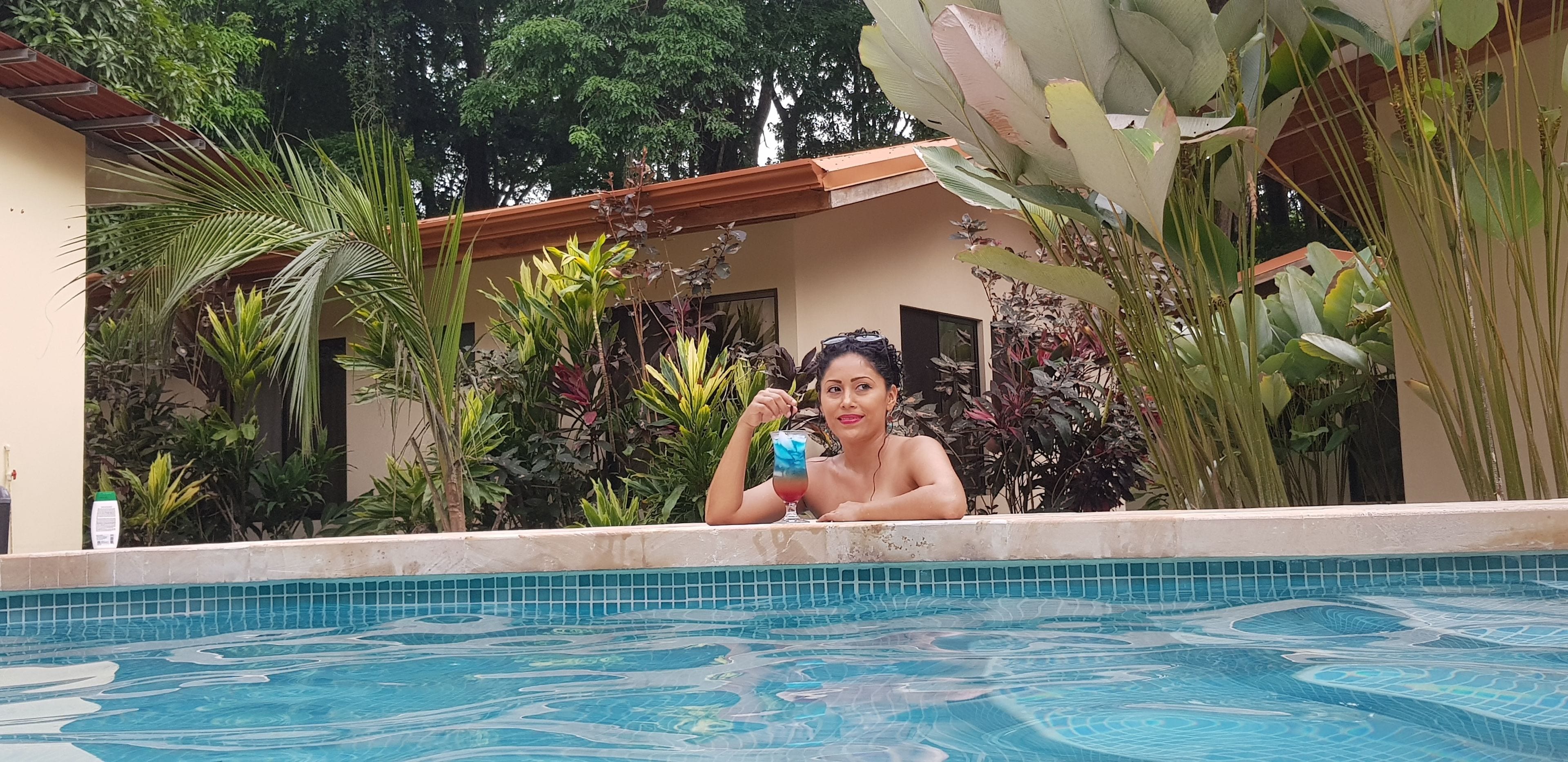 A person relaxing in a swimming pool surrounded by lush tropical plants and a building with a tiled roof.