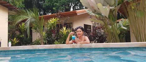 A person relaxing in a swimming pool surrounded by lush tropical plants and a building with a tiled roof.