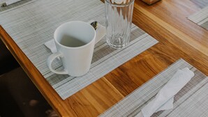 Una mesa de madera con individuales, tazas blancas y un vaso de agua.