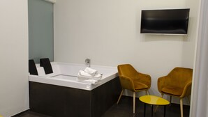 Une salle de bain moderne équipée d’une baignoire, de deux chaises, d’une petite table et d’un téléviseur à écran plat fixé au mur.