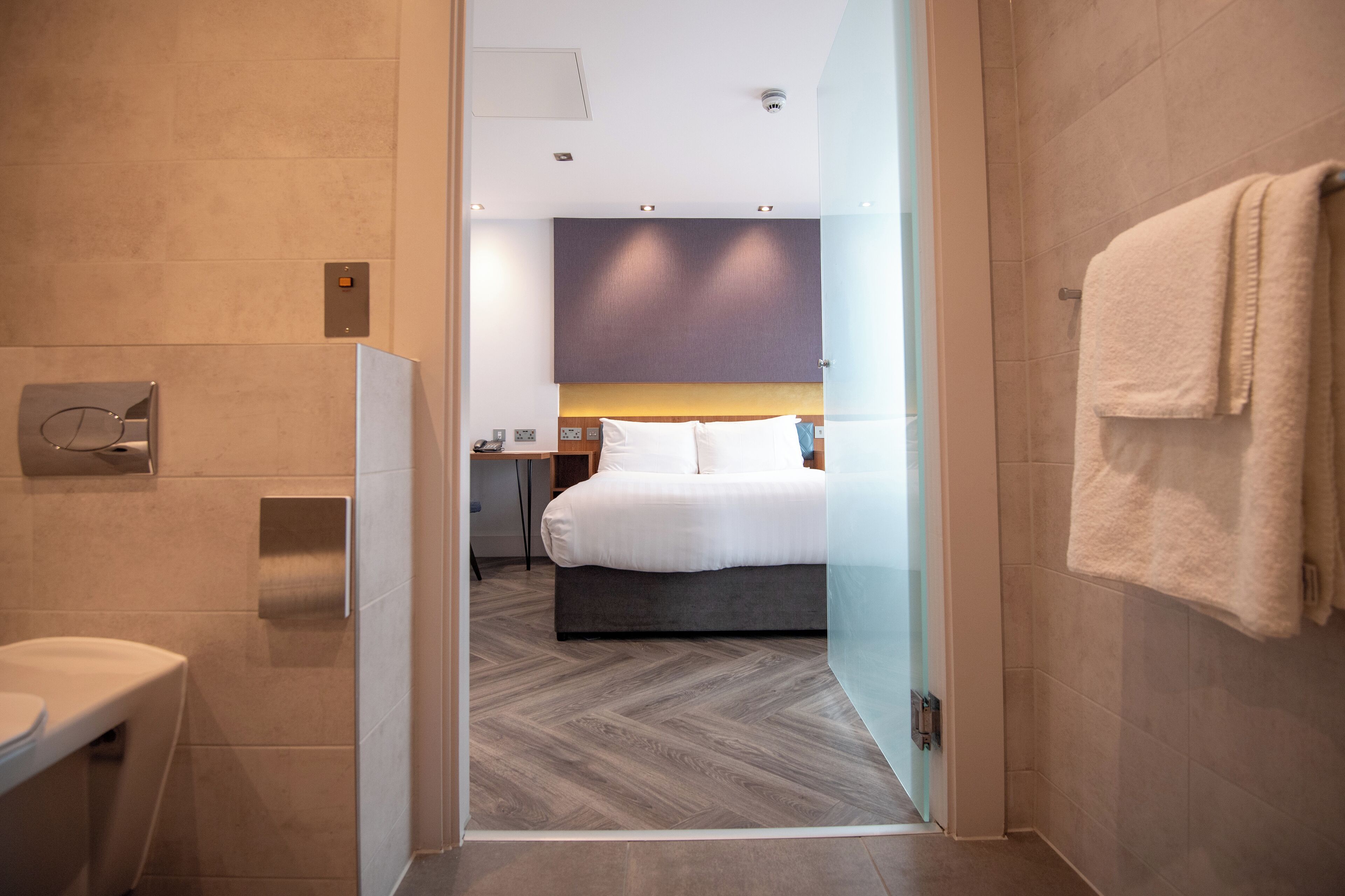 Una habitación de hotel moderna con cama, mesita de noche, baño con inodoro y perchero para toallas.