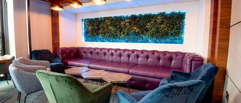 A modern lounge area with a large purple sofa, green and blue chairs, and a wooden ceiling with pendant lighting.