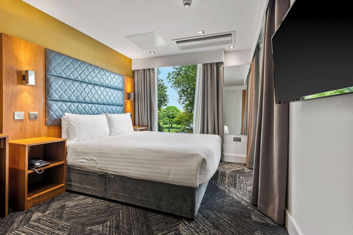 A modern hotel room with a large bed, a wooden nightstand, a built-in TV, and a view of greenery outside.