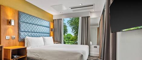 A hotel room with a large bed, a wooden nightstand, a built-in TV, and a view of the outdoors.