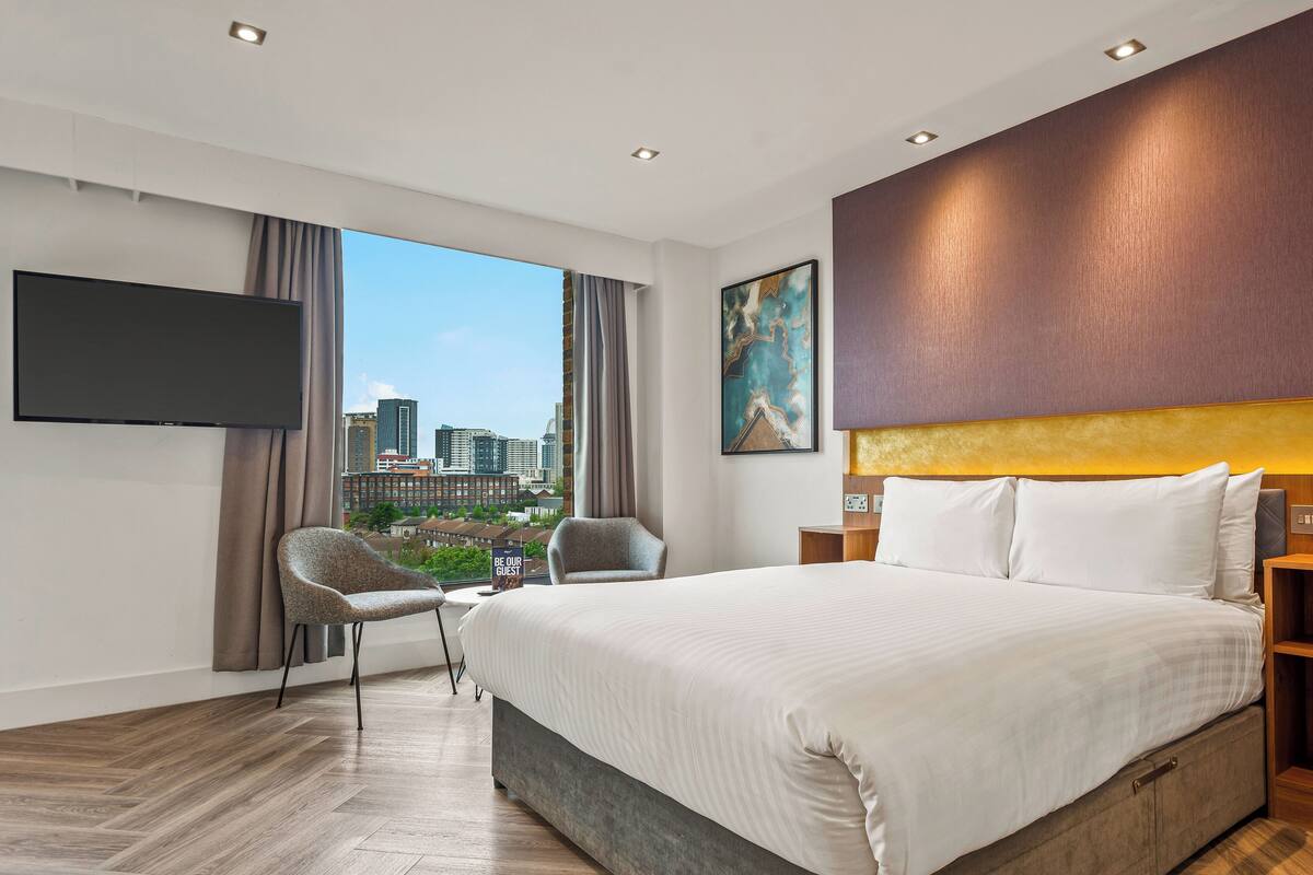 A modern hotel room with a large bed, a flat-screen TV, a city view, and a cityscape visible through the window.