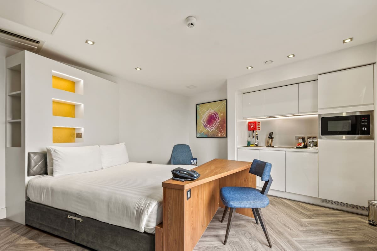 A modern hotel room with a bed, a desk, a chair, a kitchenette, and a bathroom.