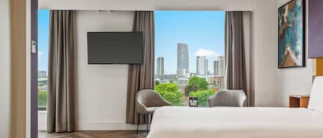 A hotel room with a large bed, a flat-screen TV, two chairs, a small table, and a city view through large windows.