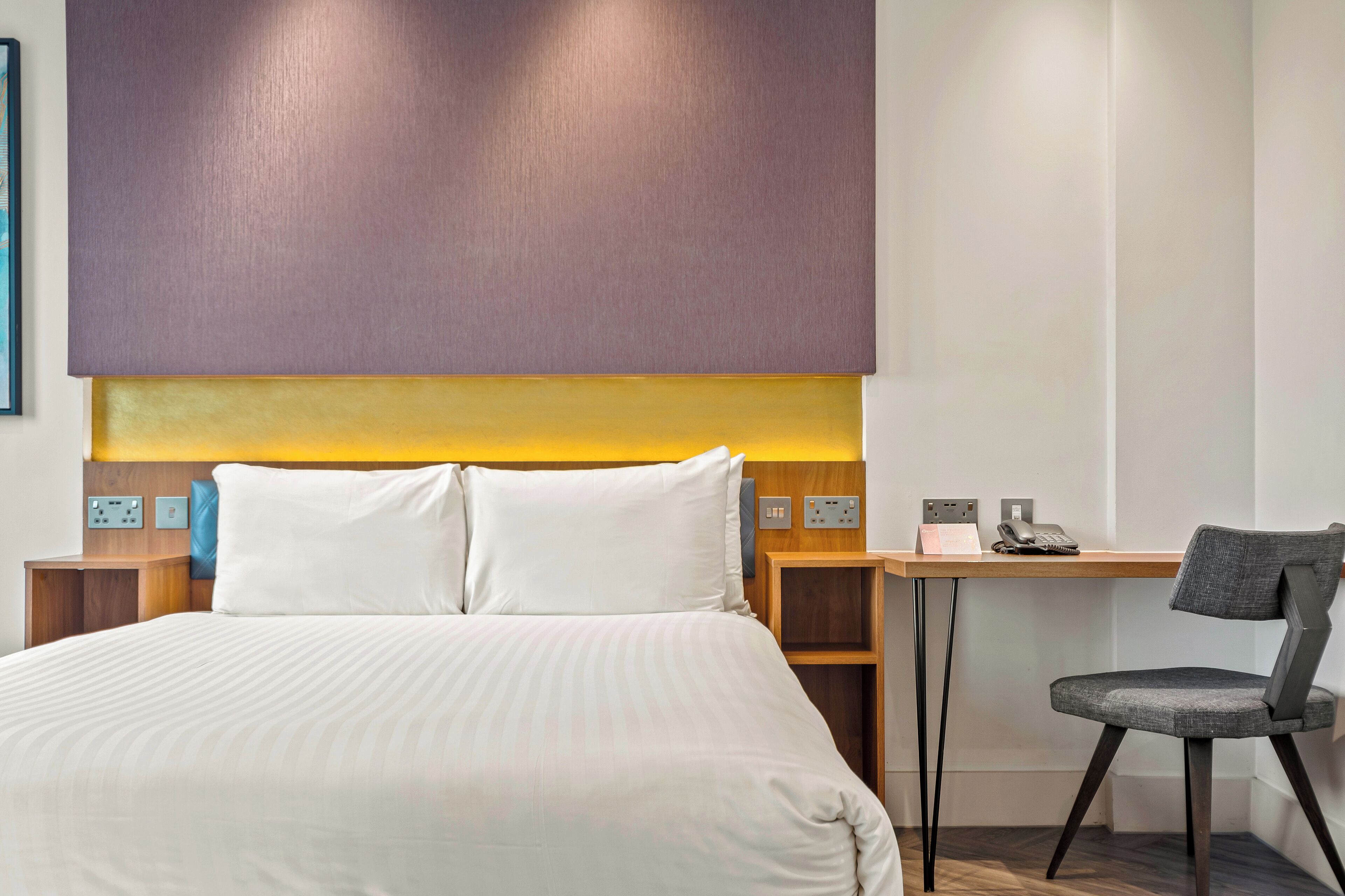 Ein Hotelzimmer mit einem großen Bett, einem Schreibtisch, einem Stuhl und einer wandmontierten Bedienoberfläche.