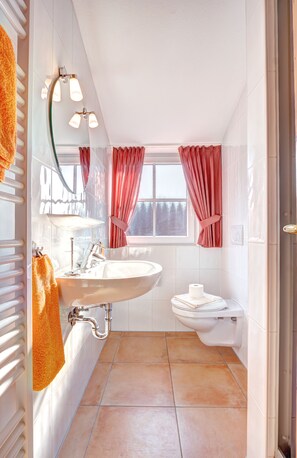 Een badkamer met een wit wastafel, een toilet en een bad. Er hangen roze gordijnen voor het raam, en een handdoek aan de radiator.
