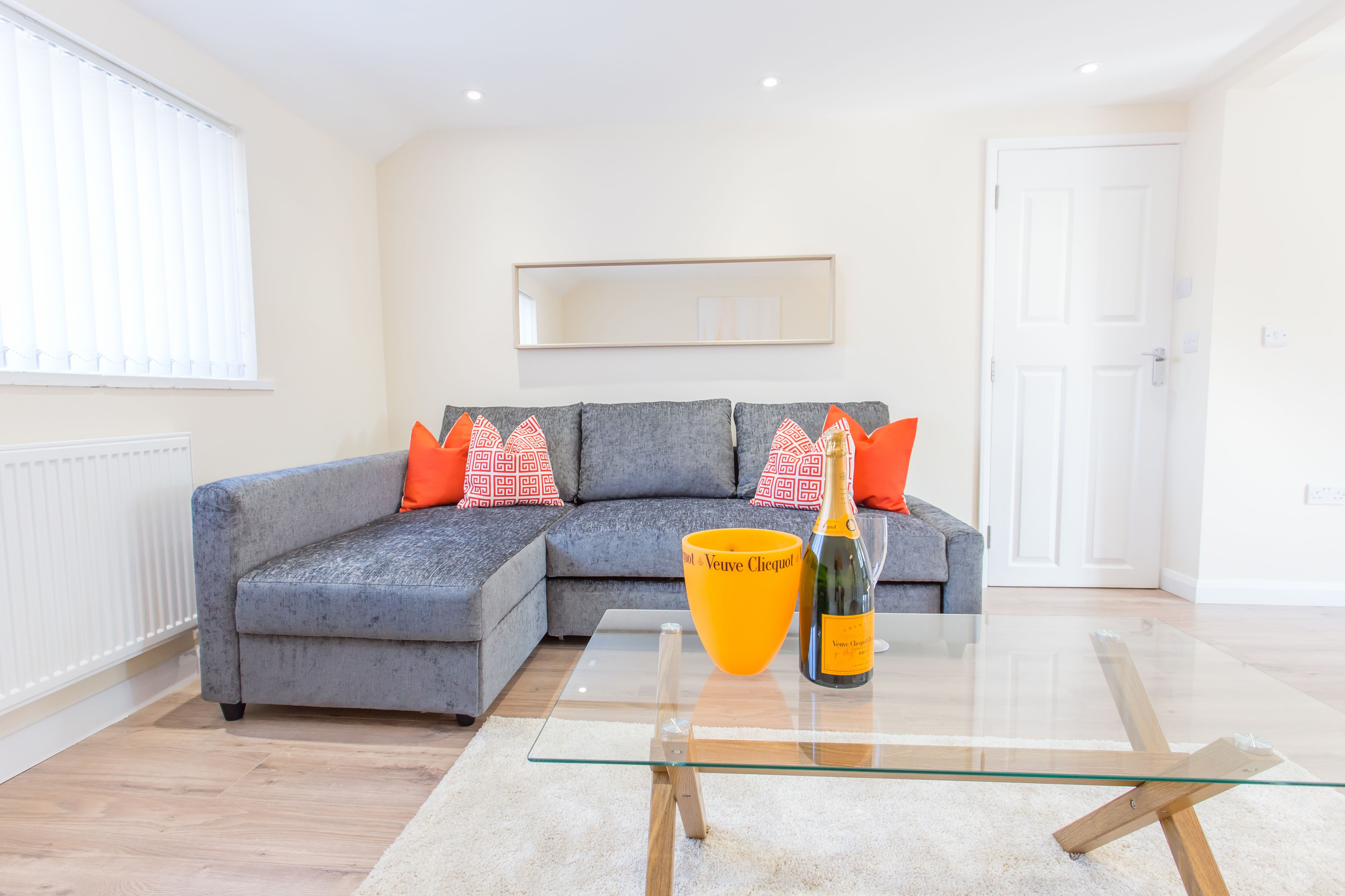 A modern living room with a grey sectional sofa, orange and white patterned cushions, a glass coffee table, and a bottle of Veuve Clicquot champagne.