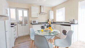 Une cuisine moderne avec une table à manger en verre, des meubles blancs, un four à micro-ondes, un lave-linge et une grande fenêtre.