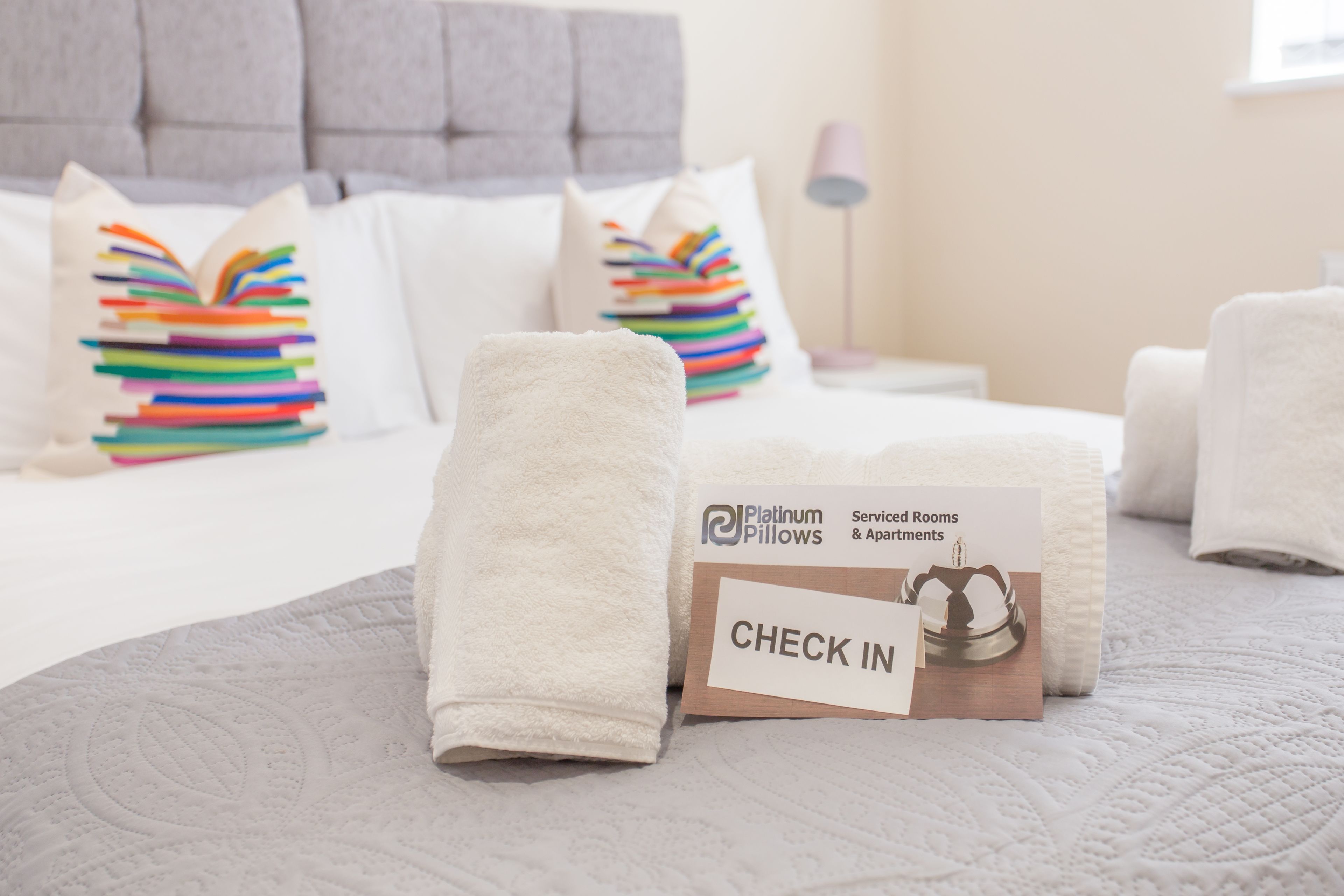 A neatly made bed with a pillow stack, a check-in card, and a bedside lamp.