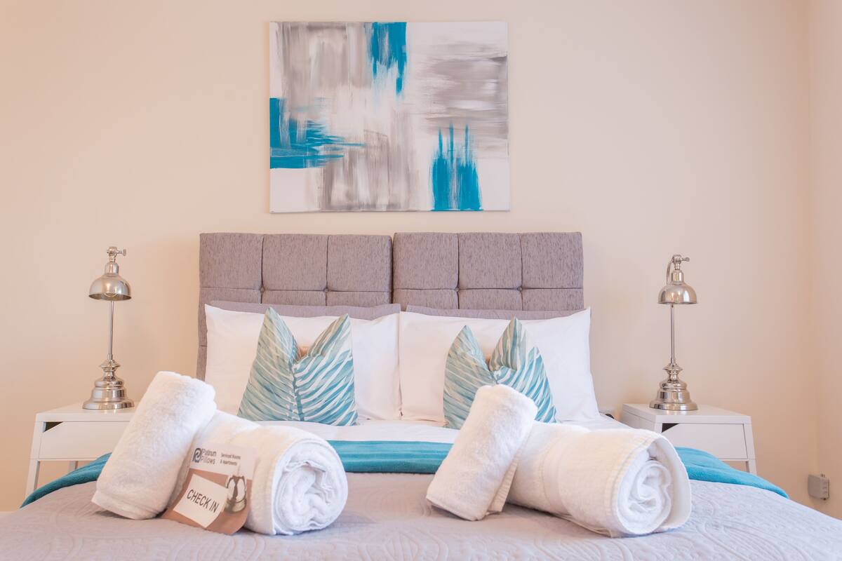 Una cama bien hecha con un cabecero gris, dos lámparas de noche y un cuadro abstracto azul y blanco encima.