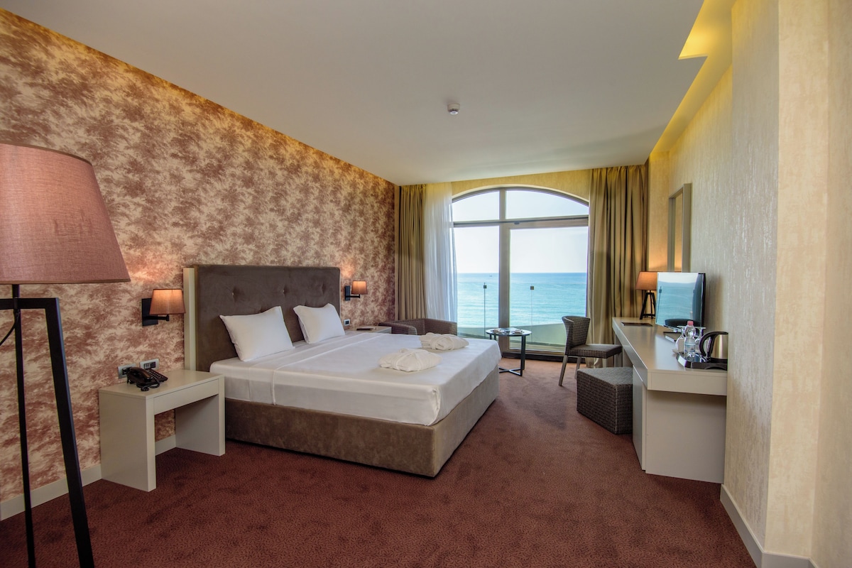 A hotel room with a large bed, a desk, a chair, a lamp, and a view of the sea.
