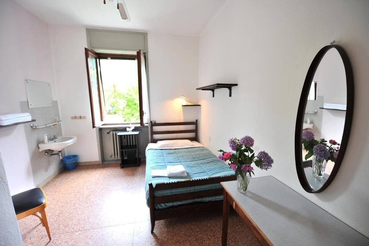 Un dormitorio con cama, mesita de noche, espejo, ventana y un pequeño escritorio con silla.