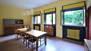 Habitación con una mesa de madera, sillas, un armario y un radiador. Se ven vistas de la naturaleza a través de las ventanas.