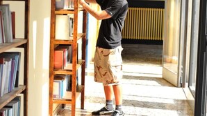 Una persona ordena libros en una estantería de madera en una habitación con suelo de baldosas y ventanas amplias.