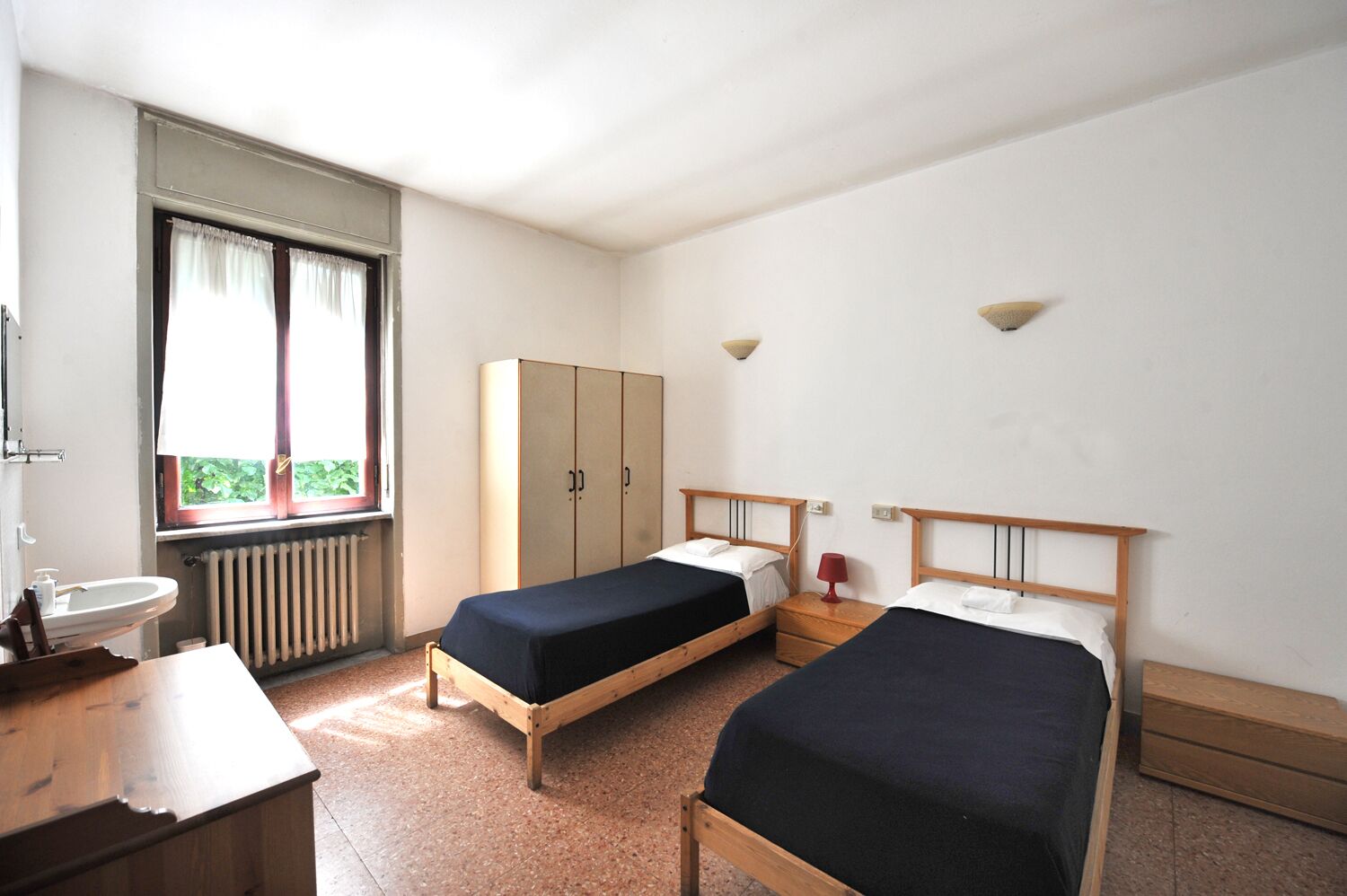 Una habitaci ón con dos camas individuales, un escritorio de madera, una ventana con cortinas, un radiador y un armario.