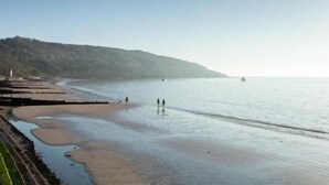 A coastal beach with a clear sky, calm sea, and a forested hill in the background. - Beachside Apartment On The Beach (Totland Bay)