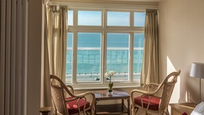 A hotel room with a large window overlooking the sea, two wicker chairs, a small table with a vase, and a bed with red pillows. - Beachside Apartment On The Beach (Totland Bay)