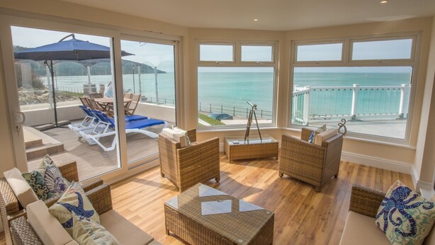 A spacious room with large windows offering a view of the sea. There are wicker furniture pieces and a glass table. - Beachside Apartment On The Beach (Totland Bay)