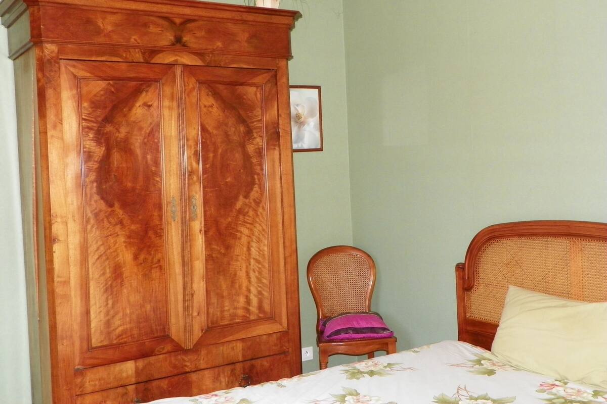 A bedroom with a wooden wardrobe, a wicker chair, a bed with a floral bedspread, and a small framed picture on the wall.