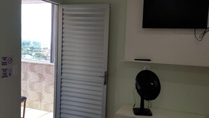 Habitación con puerta corrediza cerrada, televisor montado, ventilador de pie y balcón con baldosas y una silla.