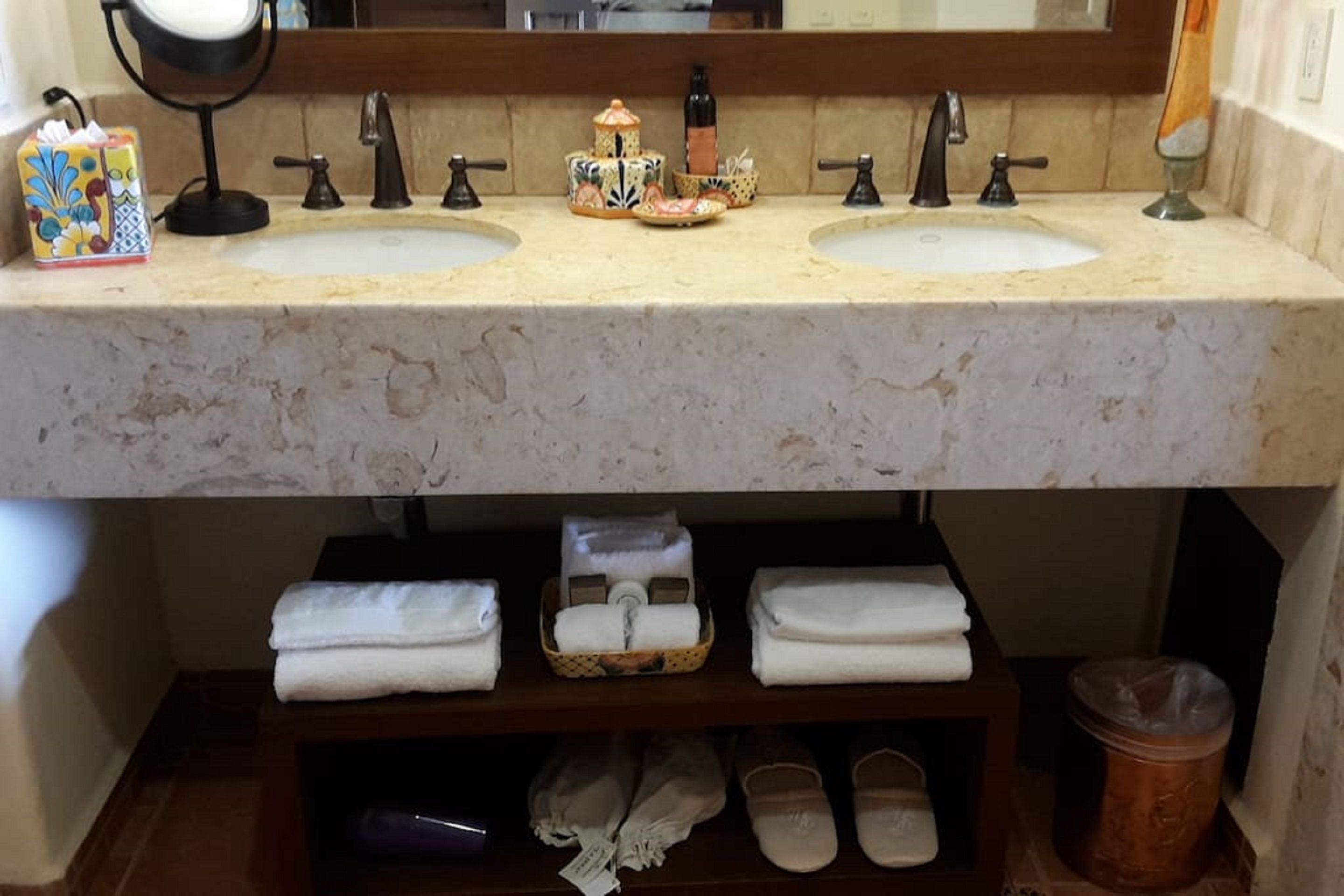 Un baño con dos lavamanos, un espejo y repisas inferiores con toallas y pantuflas.