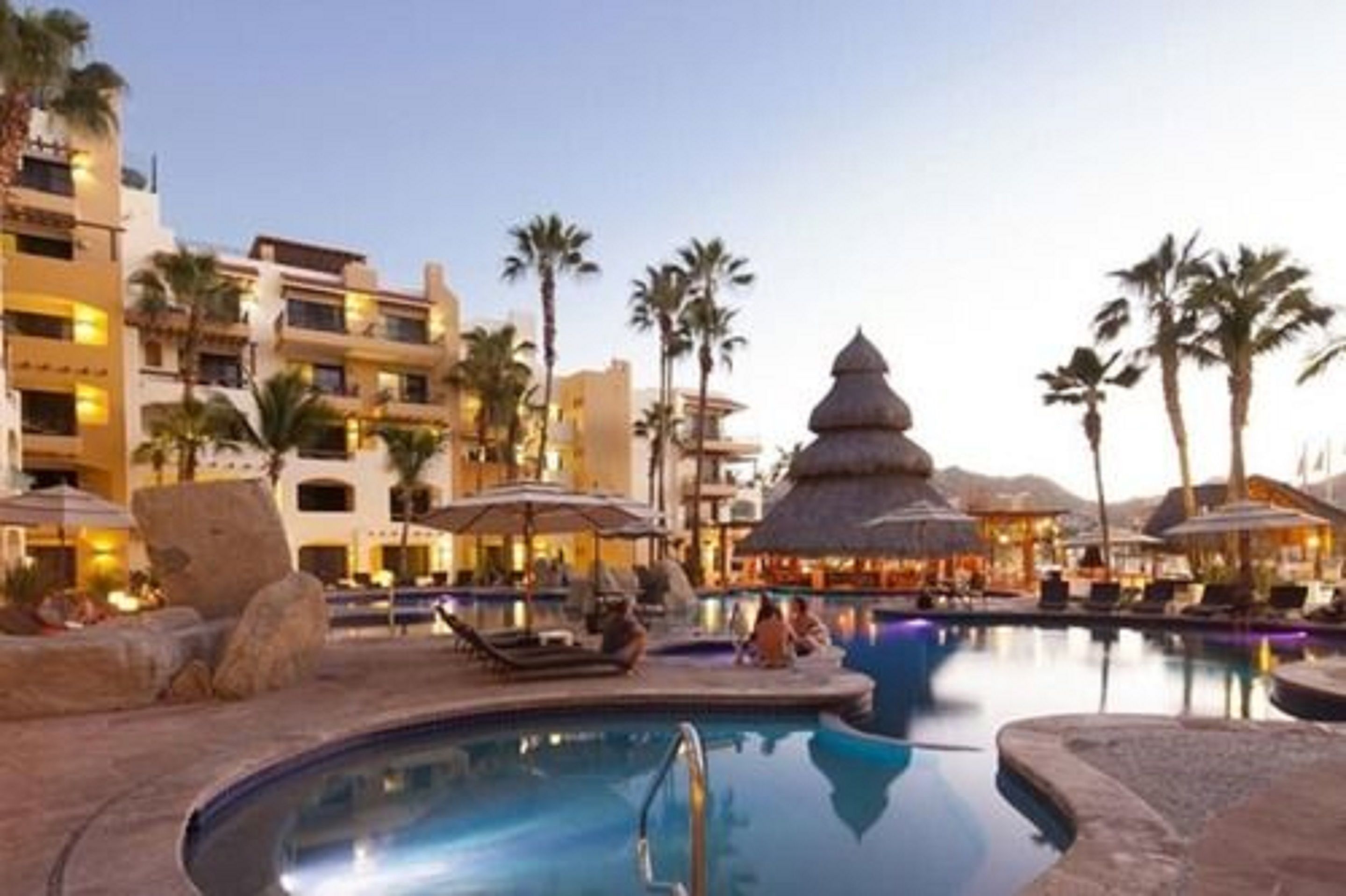 A resort pool area with lounge chairs, umbrellas, and a thatched-roof structure.