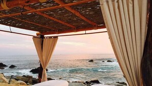 Ein Zimmer am Strand mit zwei mit weißen Laken bezogenen Betten, einer Holzbalkendecke und Blick auf den Ozean.