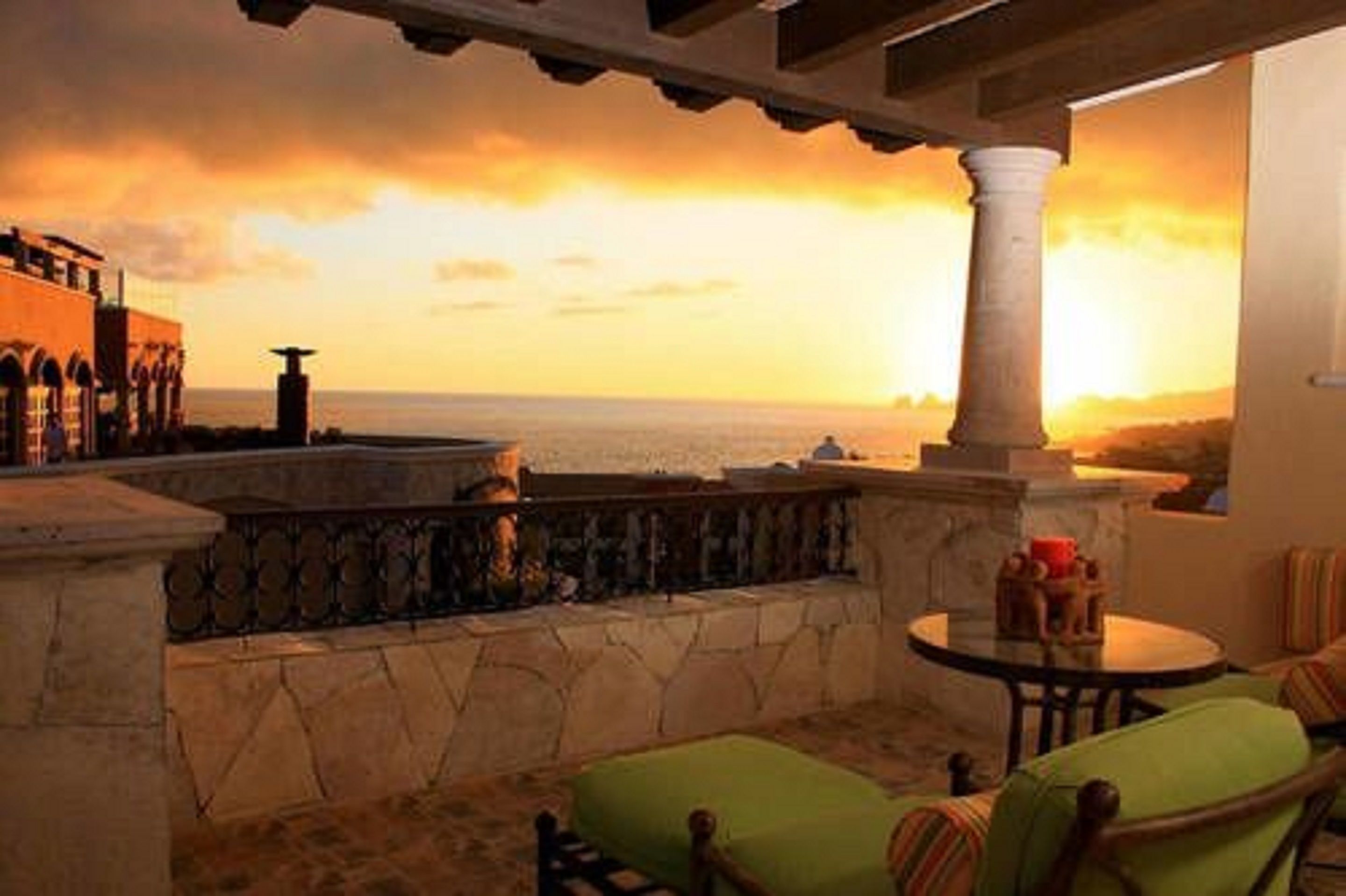 Una terrazza con vista sul mare al tramonto, ringhiere in pietra e un tavolo rotondo con sedie.