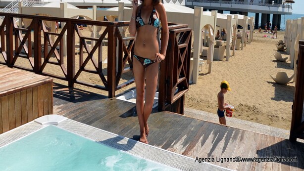 A woman in a bikini on a wooden deck with a pool, overlooking a beach with a distinctive white building with circular windows. - Villa a Lignano Riviera with air conditioning (Lignano Sabbiadoro)